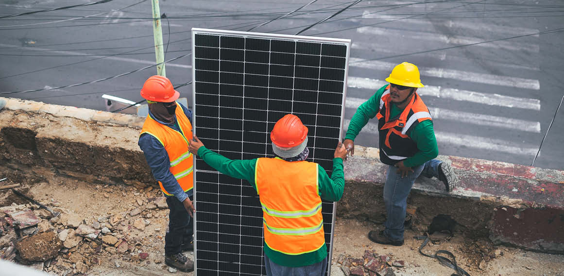 Tres obreros con casco y equipo instalando un panel solar en una obra