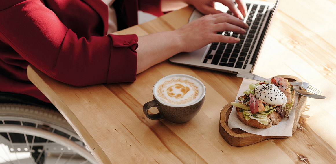 Persona escribiendo en su ordenador con una taza de café y una tostada a su derecha